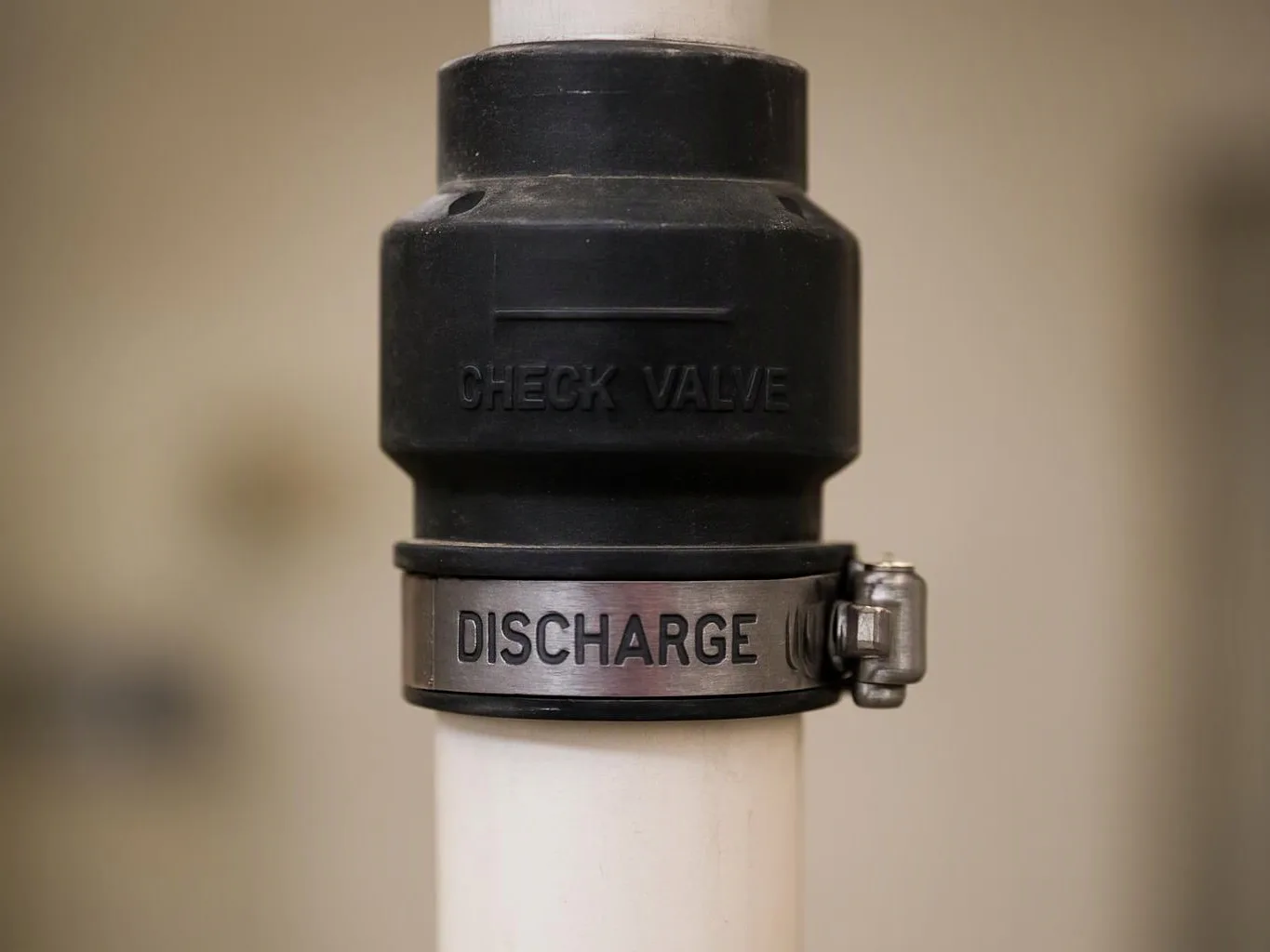 Close-up of a black check valve and metal clamp on a white pipe against a blurred beige background